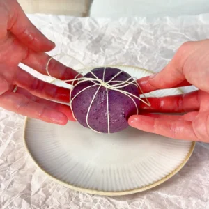Manos atando con hilo de cocina una bola de masa de bagel de arándanos morada para darle forma de calabaza