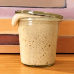 Sauerteig ansetzen: selbstgemachter Sauerteig voller Blasen, der über den Glasrand quillt und den Deckel hebt