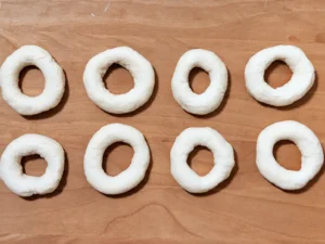 Ocho bagels de masa cruda formados con el agujero en el centro, sobre una tabla de madera